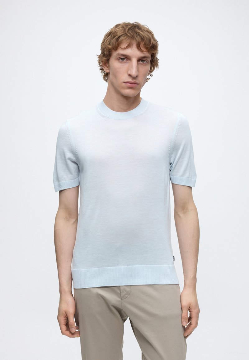Junger Mann mit kurzen lockigen Haaren, der ein hellblaues kurzärmeliges Strickshirt und beige Hosen trägt, steht vor einem schlichten weißen Hintergrund.