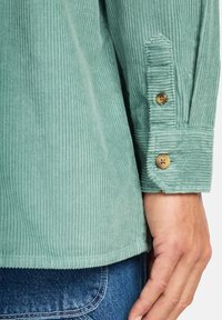 Mint green corduroy shirt with a patterned texture, featuring buttoned cuffs and a straight hem. Set against denim jeans.