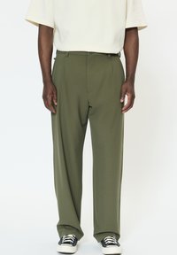 Olívazöld, sima anyagból készült bőszárú nadrág rejtett elülső zsebbel és letisztult derékpánttal, fekete sportcipővel párosítva.