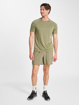 Mand, der står iført olivengrøn kortærmet skjorte og matchende shorts med sorte sneakers og hvide strømper på hvid baggrund.