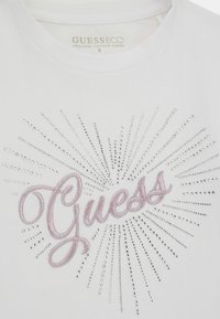Biała organiczna bawełniana koszulka z centralnym logo „Guess” w różowym haftem, ozdobiona srebrnymi kryształkami tworzącymi efekt rozbłysku gwiazdy.