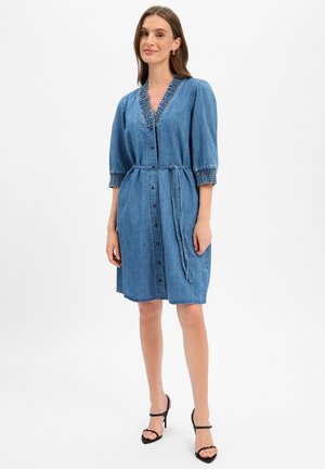 Vrouw die een blauwe denimjurk tot op de knie draagt met knopen aan de voorkant, pofmouwen, geplooide manchetten en halslijn, en een gestrikte tailleband, staand stil.