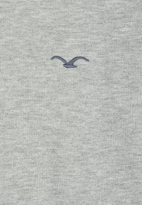 Grå stickad tröja med en liten mörkblå broderad fågelmotiv på framsidan. Mjuk textur med ett enhetligt mönster genom hela plagget.