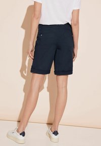 Marinblå shorts med något rullad fåll, midjehögt snitt, med fickor på sidorna och bak. Kombinerat med vita sneakers mot en ljus bakgrund.