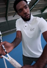 Weißes Poloshirt aus strukturiertem Stoff, mit einem Logo auf der linken Brust und schwarzen Knöpfen. Der Träger hält einen Tennisschläger auf einem blauen Platz.
