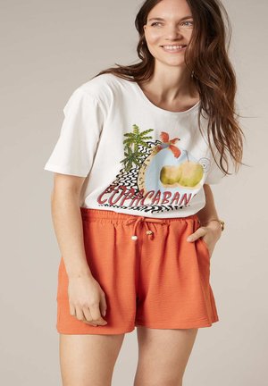 Femme souriante, portant un t-shirt blanc avec un imprimé tropical Copacabana et un short à cordon orange, main dans la poche, sur fond uni.
