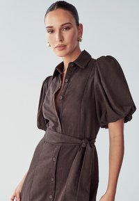 Hnědé šaty na knoflíky s nafouknutými rukávy, viditelná textilie, pás s opaskem a viditelné knoflíky vpředu.