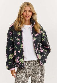 Mikina na zip z flísu s čiernym pozad�ím a ružovo-zeleným kvetinovým vzorom. Má rebrované manžety a golier, v kombinácii s nohavicami v leopardom vzore.