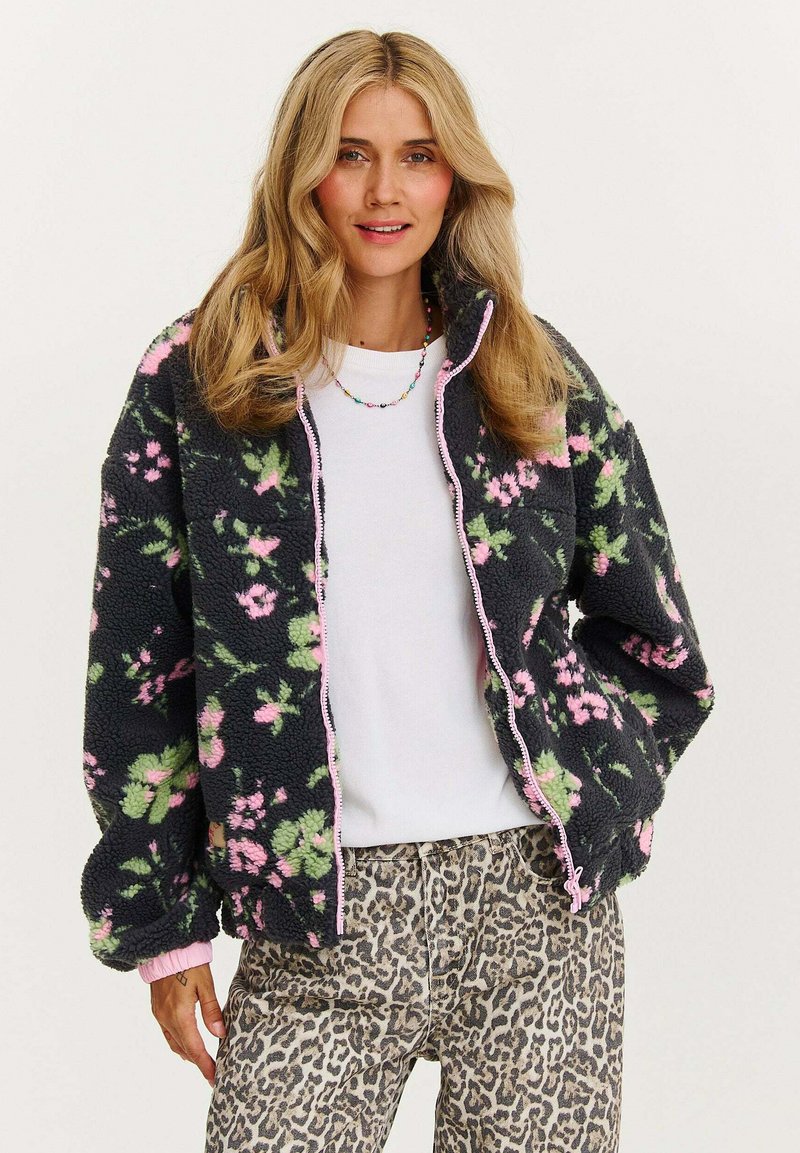 Mikina na zip z flísu s čiernym pozadím a ružovo-zeleným kvetinovým vzorom. Má rebrované manžety a golier, v kombinácii s nohavicami v leopardom vzore.