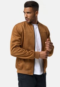 Chaqueta bomber de ante marrón con puños y dobladillo acanalados, cierre frontal con cremallera y bolsillos laterales, superpuesta sobre una camiseta blanca.