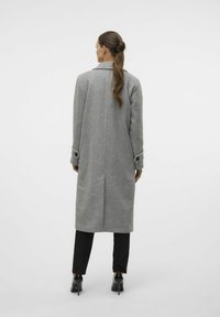 Lungo cappotto grigio in misto lana con colletto a scollo a V, polsini abbottonati e taglio dritto, abbinato a pantaloni neri e tacchi.