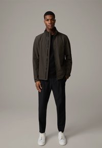 Dunkelgrüne, leichte Jacke mit hohem Kragen, Reißverschluss vorne, kombiniert mit schwarzen Hosen und weißen Sneakers. Einfaches Design, keine auffälligen Muster.