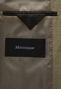 Étiquette noire "Matinique" cousue à l'intérieur de la doublure en satin beige de la veste, avec un détail en tissu noir triangulaire au-dessus de l'étiquette.
