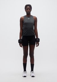 Αθλητικό άτομο στέκεται μπροστά, φορώντας μαύρο σορτς προπόνησης, σκούρο αμάνικο τοπ, βάρη αστραγάλου, μαύρες κάλτσες και λευκά αθλητικά παπούτσια.