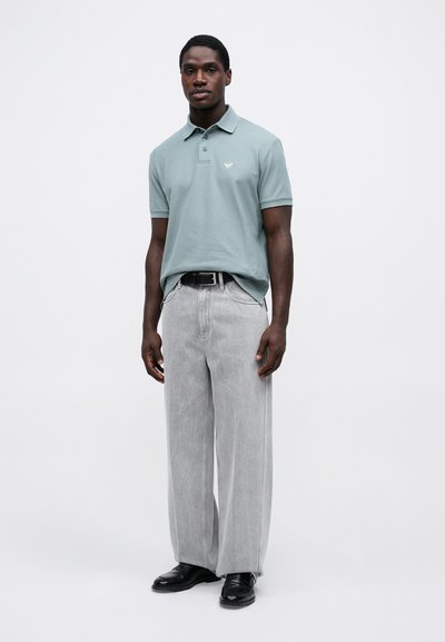 Ανοιχτό γκρι πόλο μπλουζάκι με διακριτικό λογότυπο, συνδυασμένο με φαρδιά ανοιχτό γκρι παντελόνια και μαύρα παπούτσια. Απλός, καθαρός σχεδιασμός και υφή υφάσματος.
