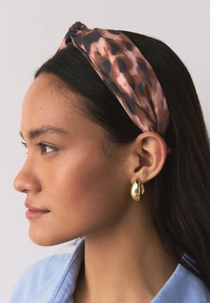 Frau mit dunklem Haar, die ein rosa-schwarz gemustertes Stirnband und einen goldenen tropfenförmigen Ohrring trägt, in einer Nahaufnahme im Profil gezeigt.