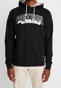 Sudadera negra con capucha forrada en gris, que presenta un gran gráfico blanco de "PETROL" en la parte frontal. Fabricada con tejido suave y de corte relajado.
