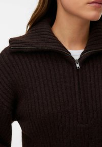 Tmavě hnědý žebrovaný pletený svetr s půlzipem u límce. Má strukturovanou tkaninu a výrazný zip u krku.