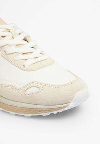 Baskets beige clair mettant en avant un mélange de matériaux lisses et texturés, des lacets blancs et une semelle à motifs avec des accents de couleur marron.