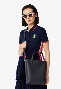 Sac à main en cuir texturé noir avec des accents rouges, double anses et bandoulière détachable. Le modèle porte un polo marine avec un emblème et des lunettes de soleil.