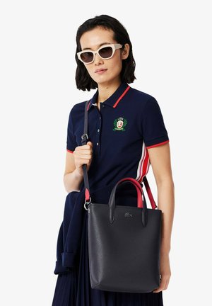 Borsa in pelle nera a texture con accenti rossi, manici doppi e tracolla removibile. Il modello indossa un polo blu navy con emblema e occhiali da sole.