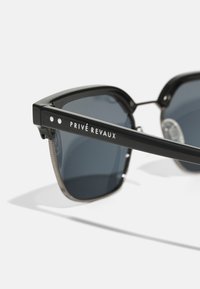 Ochelari de soare negri cu lentile colorate, având accente din metal la tâmple și inscripția "PRIVÉ REVAUX" gravată pe lateral.
