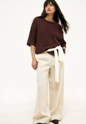 Mujer con un top de punto marrón oscuro con mangas remangadas y pantalones crema sueltos atados a la cintura con una faja blanca sobre un fondo blanco.