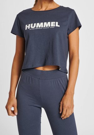 Kobieta w pasującym granatowym krótkim topie z logo "HUMMEL" i legginsach z wysokim stanem, ramiona swobodnie opuszczone wzdłuż ciała, neutralne tło.