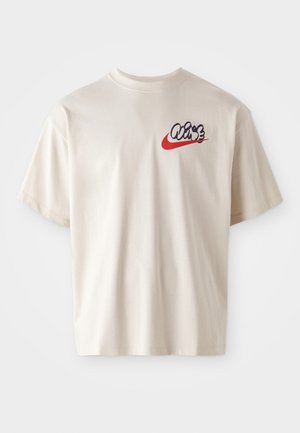 T-shirt em algodão bege com mangas curtas, apresentando um gráfico colorido no logo na área do peito esquerdo, combinando acentos azuis e vermelhos.