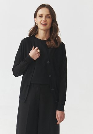 Cardigan nero con chiusura a bottoni sul davanti, maniche lunghe e polsini a costine, indossato sopra un top coordinato e abbinato a pantaloni neri.