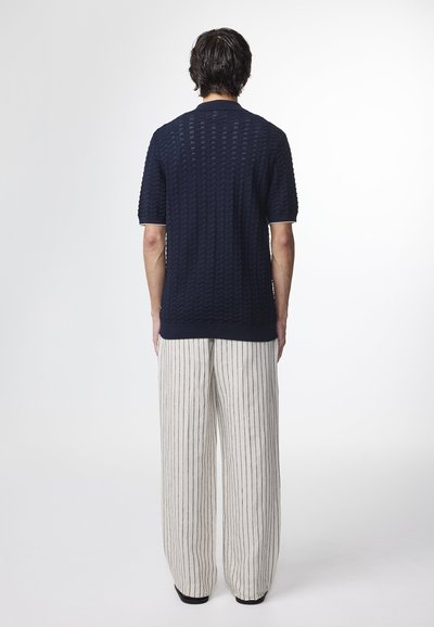 Marineblå polo t-shirt med strukturerede bølge mønstre, korte ærmer og kontrastfarvet krave; parret med løse, stripede linnedbukser i neutrale toner.