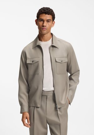 Jeune homme portant une veste zippée beige clair sur une chemise blanche avec un pantalon beige assorti, debout devant un fond blanc uni.