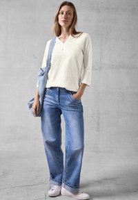 Helles beiges Langarmhemd, lässig geschnittene verwaschene blaue Jeans, weiße Sneakers und eine hellblaue Umhängetasche vor einem grauen Hintergrund.