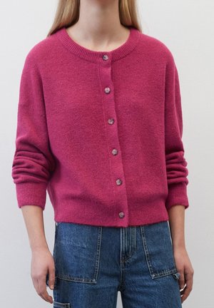 Femme portant un cardigan rose vif boutonné à manches longues et un jean en denim bleu taille haute, sur un fond clair uni.