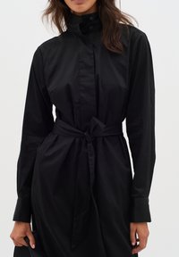 Svart långärmad klänning med hög krage, knytmidja och knappuppknäppning. Tillverkad i ett mjukt tyg med en elegant, minimalistisk design.