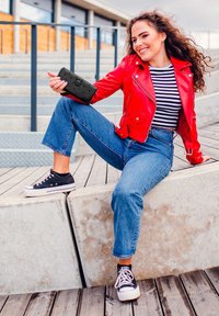 Black wallet with textured surface held in a hand. Red leather jacket, striped shirt, and blue denim wide-leg jeans. Black sneakers.