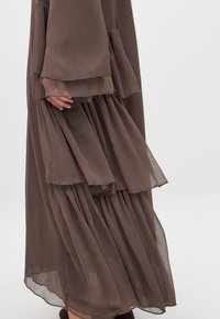Abito marrone a strati, plissettato, realizzato in tessuto trasparente, con maniche arricciate e orlo irregolare. La texture appare morbida e leggera.