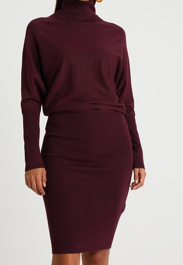 Donna che indossa un vestito a maglia attillato con maniche lunghe e collo alto di colore bordeaux, su uno sfondo chiaro e uniforme.