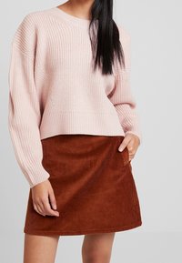 Hellrosa gestrickter Pullover mit weiten Ärmeln, kombiniert mit einem rostfarbenen Cordrock mit geradem Saum und seitlichen Taschen.