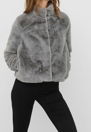 Femme portant une veste en fausse fourrure grise douce avec un col montant et un pantalon noir ajusté, debout les mains dans les poches de la veste.