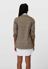 Hnědý kostkovaný blazer s přiléhavým střihem, ohrnutými rukávy a upravenou siluetou, nošený přes bílé šaty košového typu.