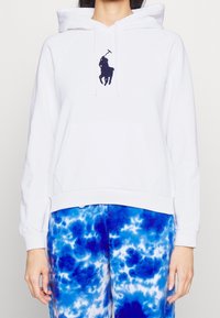 Vit luvtröja med en framficka, med en marinblå broderad logotyp. Parat med blåa tie-dye byxor som betonar livliga färger.