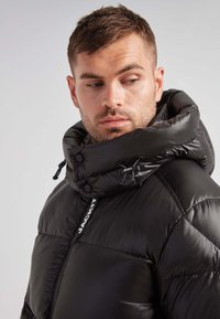 Giacca a vento nera con cappuccio alto e regolabile, texture lucida e dettagli con logo bianco. Presenta cuciture in evidenza e chiusura con zip.