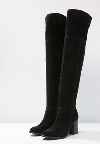 Bottes noires au-dessus du genou en daim avec un talon block. Présente une silhouette ajustée et une texture lisse avec un détail de fermeture éclair sur le côté.