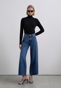 Donna con dolcevita nero, jeans azzurri a gamba ampia, sandali argentati con tacco alto e occhiali da sole neri in piedi davanti a uno sfondo uniforme.