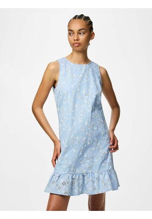 Žena v svetlomodrých kvetovaných šatách bez rukávov s volánovým lemom, stojaca s jednou rukou v páse, pozerajúca sa do strany.