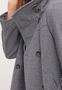 Abrigo de lana gris con cuello alto, cierre de botones doble y bolsillos laterales. Textura suave con botones oscuros y detalles de costura.