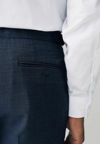 Pantalones texturizados en azul marino con un bolsillo trasero que cuenta con un botón, combinados con una camisa blanca de manga larga. Detalles sutiles en la trama de la tela.