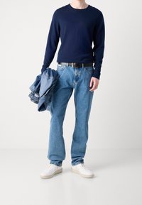 Camicia a maniche lunghe blu navy, jeans in denim blu chiaro, cintura nera, sneakers bianche. Tiene una giacca in denim. Outfit casual con vestibilità rilassata.