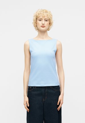 Personne aux cheveux blonds courts et bouclés, portant un haut sans manches bleu clair et un jean large foncé, debout devant un fond blanc uni.